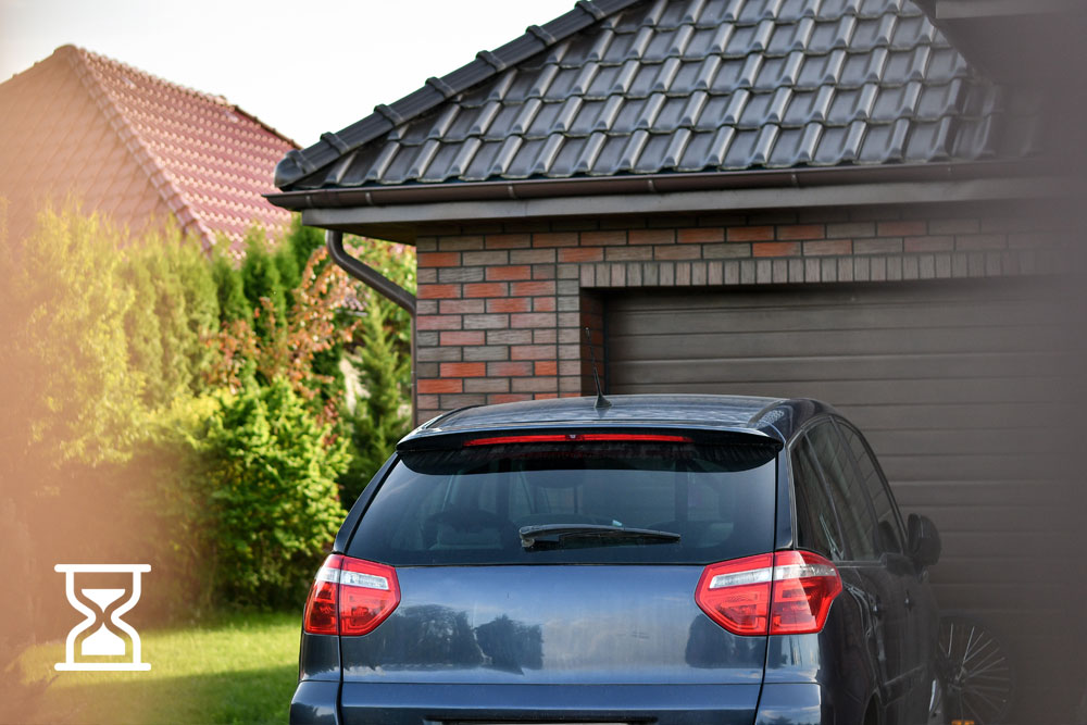 A small blue sedan is parked in front of a brick house with a brown garage door. A small, white hourglass icon sits in the lower left hand of the image.