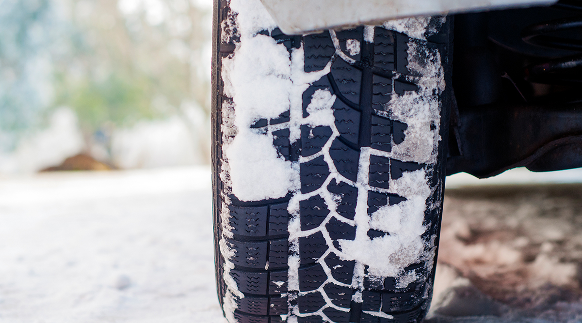 Close up of a vehicle's rear tire with snow on the tread; in the background is more snow and trees